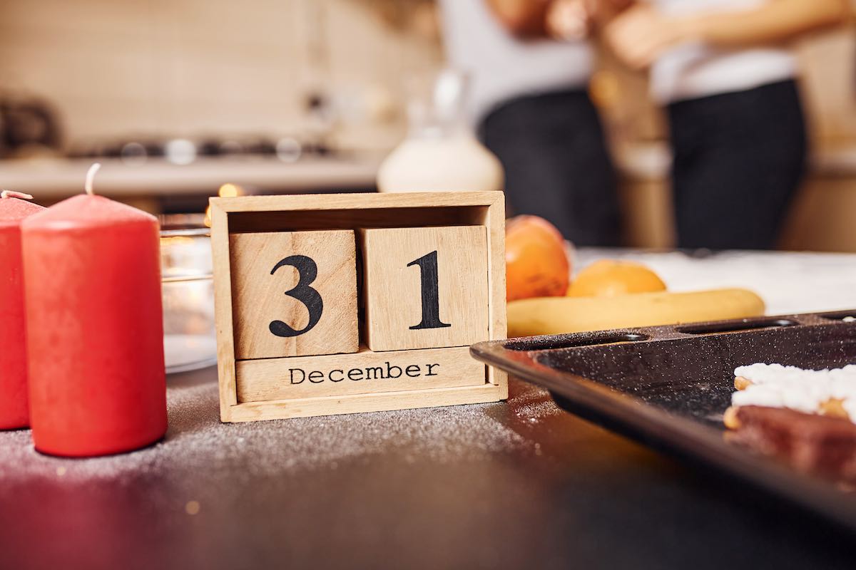 Wooden calendar with 31 december writen on it. Candles and food on the table. People at background